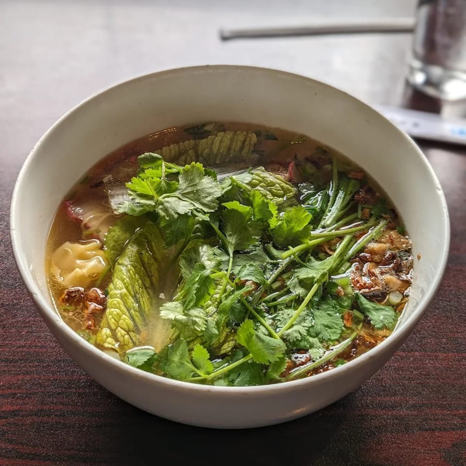 A white bowl of Vietnamese wonton soup with fresh cilantro, lettuce, and fried garlic.